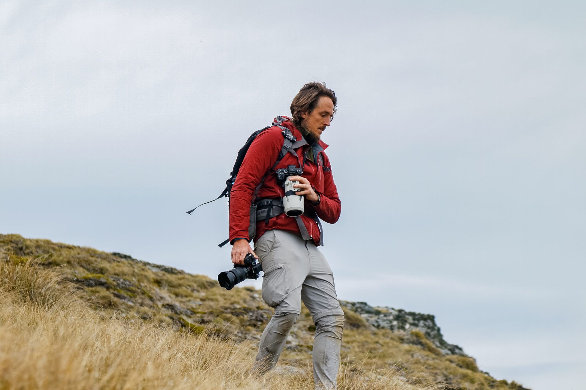 "Outdoor adventure photographer trekking through New Zealand's alpine terrain, carrying professional camera gear. Showcasing rugged exploration and on-location photography in Queenstown."