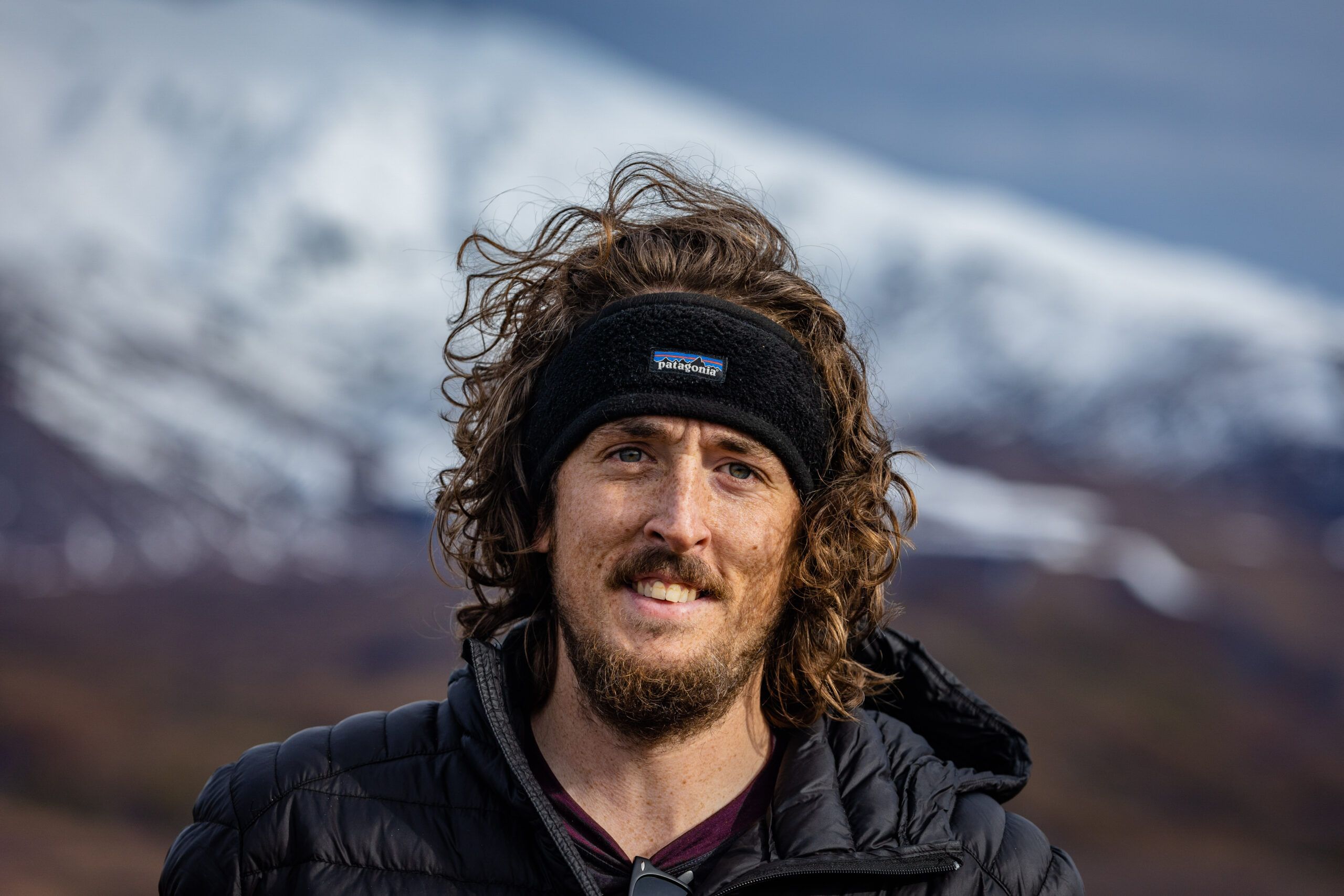 "Adventure photographer outdoors in New Zealand with snow-capped mountains in the background, wearing a Patagonia headband and a down jacket. Capturing the essence of adventure and nature in Queenstown."